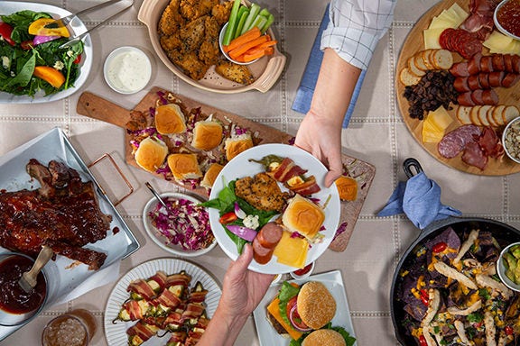 Overhead view of a shared table with fried chicken, sliders, salads, sausages, ribs, and sides as hands place a filled plate