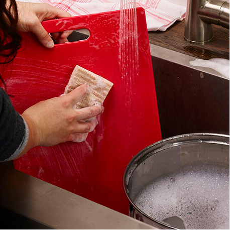 Red cutting board being cleaned in kitchen sink with sponge